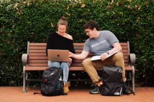 Two people sitting on a bench, looking at a laptop, with backpacks on the ground.